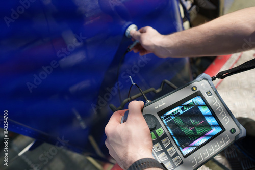 An aviation technician conducting ultrasonic testing on a blue aircraft rudder using portable NDT equipment with a digital display for defect detection, structural integrity inspection and maintenance