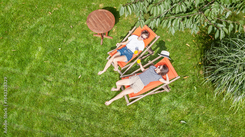 Naklejka premium Young girls relax in summer garden in sunbed deckchairs on grass, women friends have fun outdoors in green park on weekend, aerial drone view from above