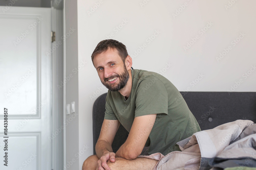 Fototapeta premium Adult man sitting on bed at home and smiling at camera, relaxed casual lifestyle in cozy bedroom interior, morning routine and everyday life concept, natural light and authentic domestic atmosphere