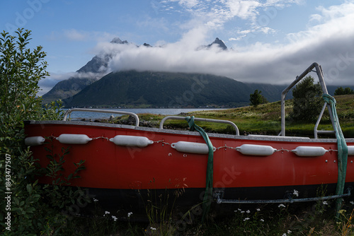 Red fishing boat on the green shore with fjord behind and cloudy covered mountains at Lofoten Island during summer season. Norway
