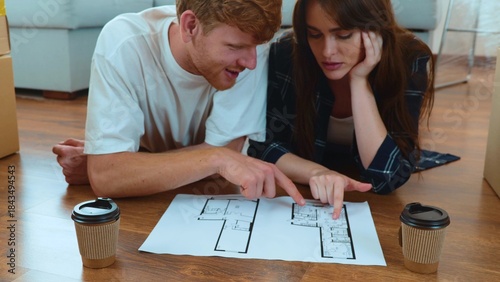 Cute young Caucasian boyfriend and girlfriend lying on the floor in new apartment with sofa and many boxes looking at plan documentation and talking. New own home. Relocation. Moving in