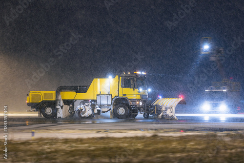 Special snow plow truck on the move along airport runway during heavy snowfall. Scene shows winter maintenance in harsh weather at night.