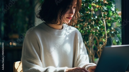 窓辺でパソコン作業に集中する女性の姿。仕事や学習シーンに適したイメージ素材。