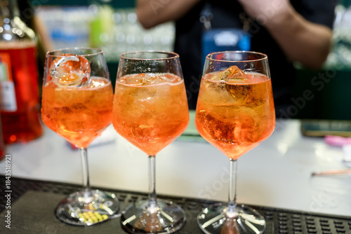 Three stemmed wine glasses filled with orange Aperol Spritz cocktails and ice cubes rest on a bar counter