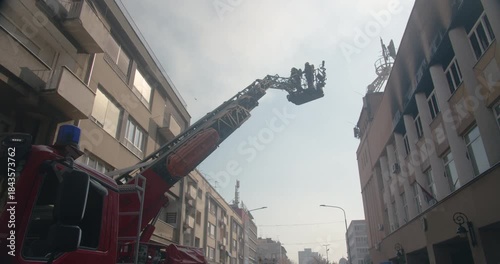 Silhouette of firefighters in a bucket on the ladder of a fire truck lifting into the air into the roof of a burning building