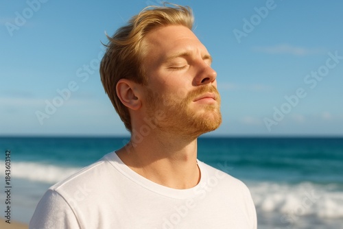 Relaxed handsome man enjoying sun at beach, eyes closed. Concept of vacation, mindfulness, and wellbeing on sunny day by ocean. Guy taking deep breath, appreciating serene sea view and clear sky