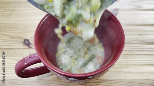 Closeup of thick homemade mixed vegetable soup, hot and steaming, pouring from a pan or pot into a large bowl, ready to eat.