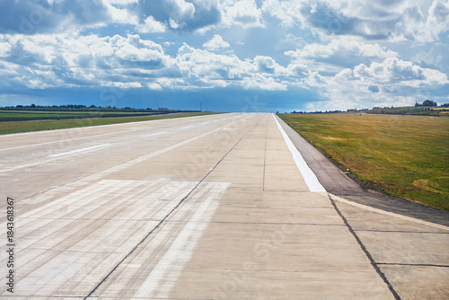 Wide concrete runway stretches into distance beneath a partly cloudy sky, flanked by grassy fields. White markings guide aircraft takeoff and landing, with distant buildings lining the horizon