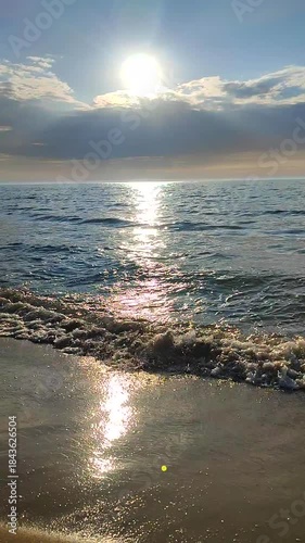 Seascape on sunny summer day. Sea waves rolling on sand of sandy beach on seashore. Waves of calm sea, sand, blue sky with clouds and horizon line. Sun shining from behind rainy clouds. Vertical