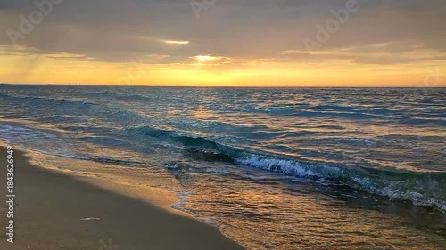 Sea waves with blue transparent water on sand of seashore on sunset and sunrise. Yellow reflection of sunlight on surface of sea waves. Sky with dark rain clouds, horizon line and sea waves on sand.