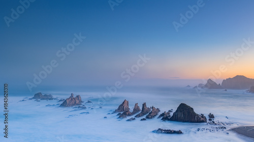 Gueirua Beach in Asturias at dawn, bathed in the first light of day. A peaceful coastal scene with dramatic rocks, soft colors, and a calm atmosphere that captures the beauty of early morning.