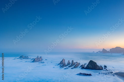 Gueirua Beach in Asturias at dawn, bathed in the first light of day. A peaceful coastal scene with dramatic rocks, soft colors, and a calm atmosphere that captures the beauty of early morning.