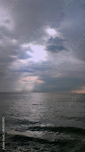 Rain in sea. Rain falling in sea. Cloudy, rainy, stormy weather at sea. Dark rainy clouds over water. Sea, sky, clouds and horizon line. Seascape. Landscape. The sun shining through clouds Vertical