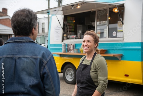 A smiling woman in an apron stands in front of a food truck, talking to a customer.