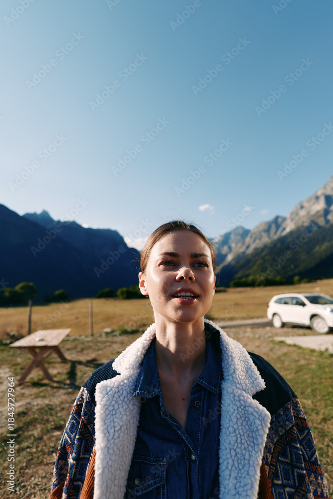Fototapeta premium woman mountains outdoor car portrait nature: young female traveler in a shearling denim jacket stands in an alpine meadow by a picnic bench and parked vehicle under clear blue sky.
