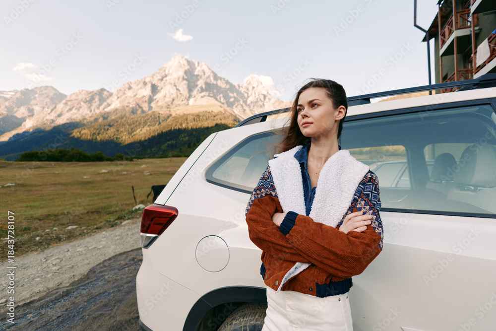 Fototapeta premium Woman leaning against white car in mountains landscape, portrait of a confident traveler on a roadtrip. Travel adventure outdoors scene with warm jacket, arms crossed and serene scenery.