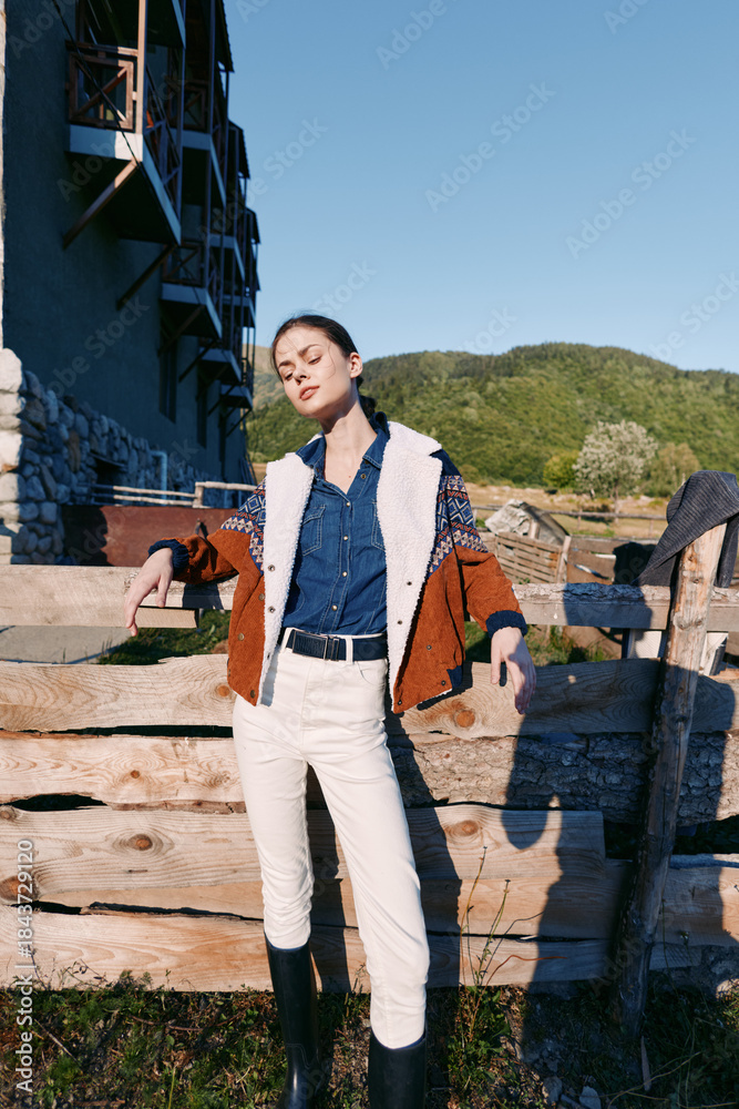 Fototapeta premium Woman leans on wooden fence in countryside with denim jacket, boots and white pants, posing relaxed against mountains and sunlight for fashion portrait, rural lifestyle and travel.