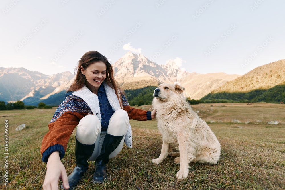 Fototapeta premium Woman, dog, mountains, meadow, outdoor, pet, happy, nature — Young woman crouching and smiling beside fluffy dog on grassy meadow with mountain backdrop, enjoying hiking and pet companionship.