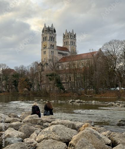 Two women sitting and talking on the shores of the Isar River  in Munich Germany on a winter day with St. Maximilian Catholic Church in the background.