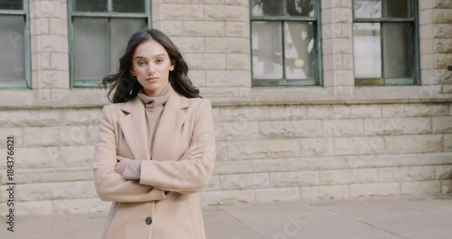 A brunette businesswoman crossing her arms and looking at the camera, with a modern building in the background and a large black car approaching behind her as she hurries.