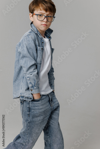 Caucasian boy wearing denim jacket and jeans poses confidently against a neutral background, showcasing a casual style with a modern touch and youthful charm