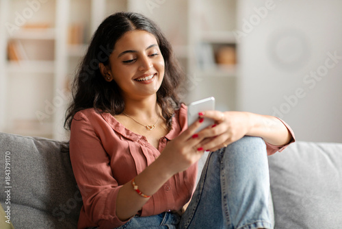 Happy Indian lady texting on smartphone, browsing social networks or shopping online while relaxing on couch at home in living room