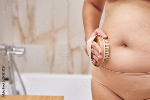 Adult Caucasian female dry brushing stomach in bathroom, wellness routine, natural body texture, self care practice and body acceptance concept.