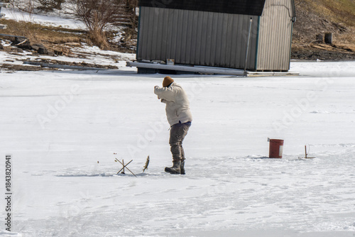 A winter scene capturing a person engaged in ice fishing on a frozen lake. Dressed in warm clothing, the individual bends over near a hole in the ice, handling a fishing rod or tip-up device. 