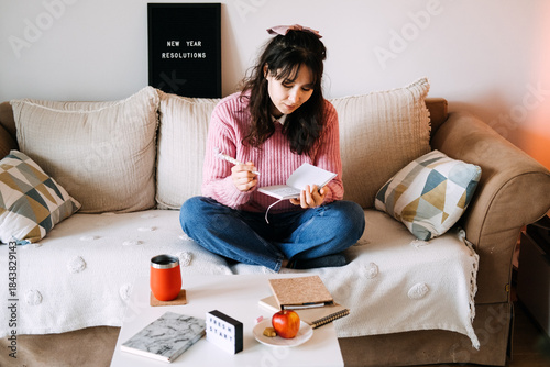 Young adult woman sits cross-legged on sofa writing in small notebook next to letter board displaying NEW YEAR RESOLUTIONS. Goal setting, New Year resolutions, mental health, healthy lifestyle