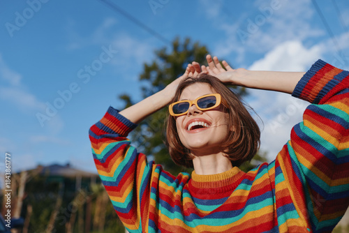 Woman smile sunglasses sweater happy portrait outdoors sunshine in colorful striped jumper, cheerful young adult enjoying a sunny day with hands raised and looking up at sky.