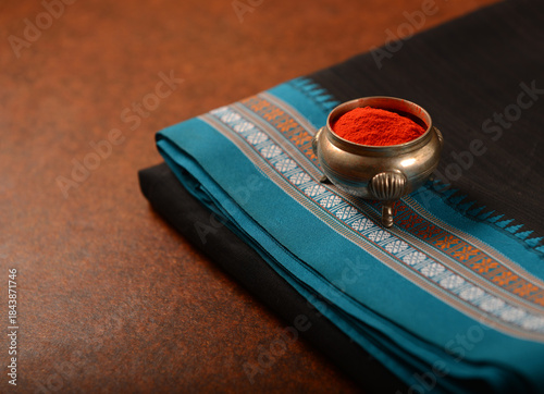 An ornate silver bowl filled with vibrant red Kumkum powder rests on a folded black saree with detailed teal boarder. A Makar sankranti festive objects and background.
