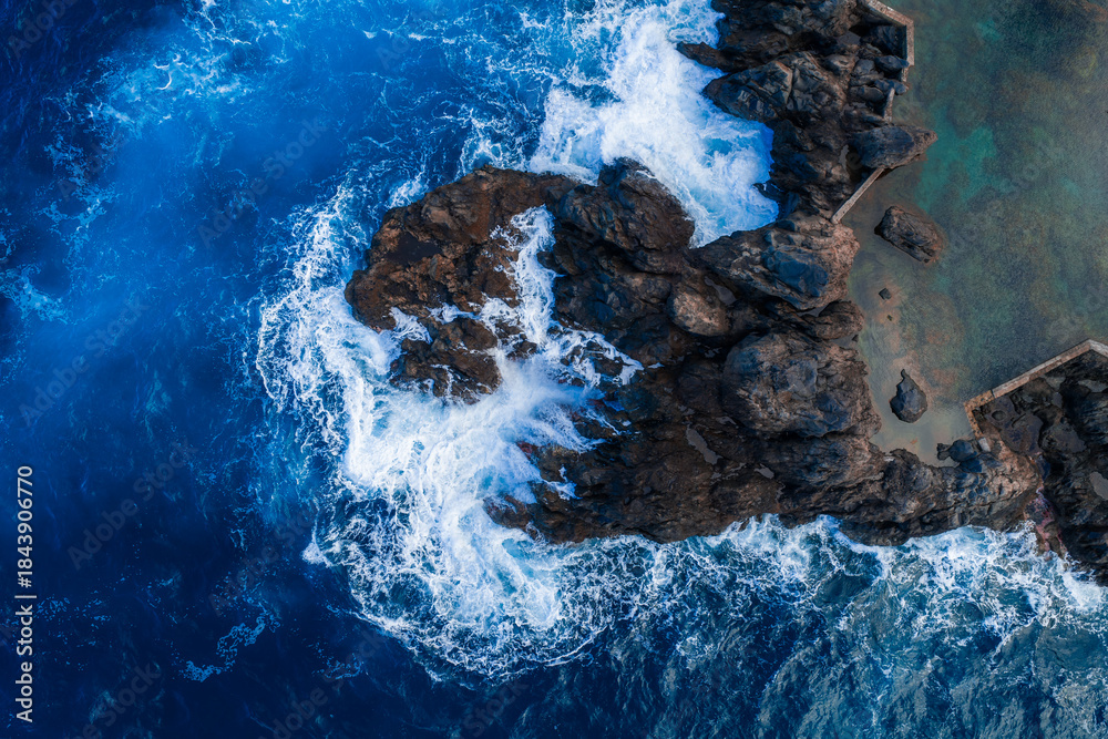 Obraz premium Aerial waves hit jagged volcanic rocks by a stone edged sea pool at Madeira, Portugal. Turquoise to deep blue water churns in bright daylight with high contrast.