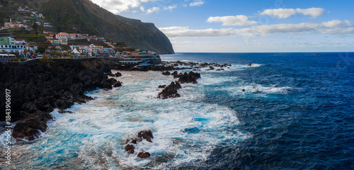 Fotografija Daylight panoramic view of Porto Moniz, Madeira, with waves striking black lava rocks, whitewashed houses on a slope, a towering headland, and Atlantic waters under clouds