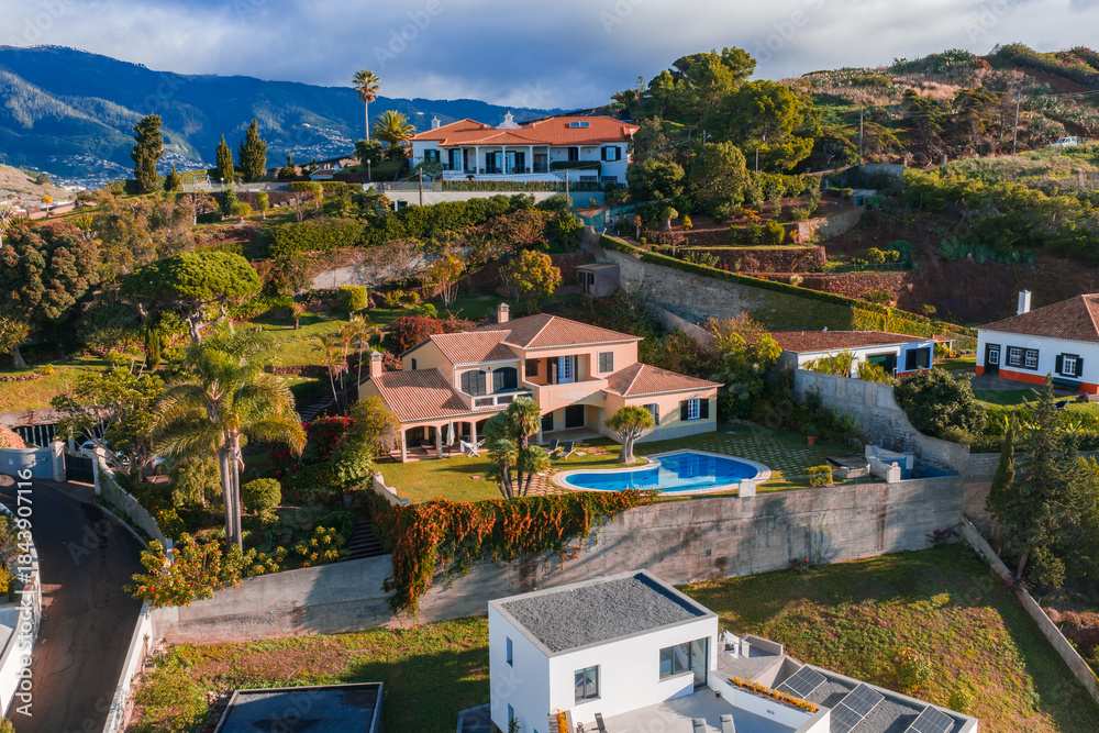 Fototapeta premium Aerial view of terraced villas on Madeira Island, Portugal, centered on a peach villa with arched verandas and a blue pool, warm light, palms, and winding roads.