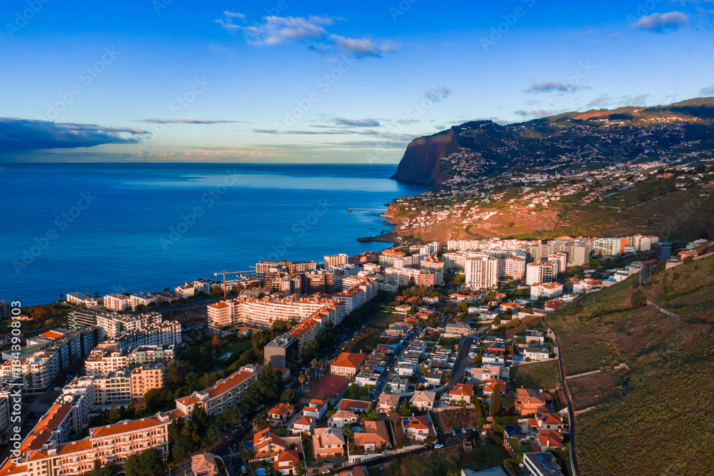 Fototapeta premium Aerial view of Funchal, Madeira, Portugal shows whitewashed blocks and hotels by the Atlantic, Cabo Girao in distance, warm late afternoon light, clear blue sky.
