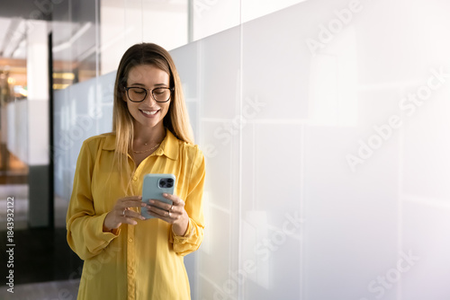 Positive stylish young professional woman in glasses typing on smartphone in office corridor, browsing social media, Internet on work break, enjoying online communication, wireless technology
