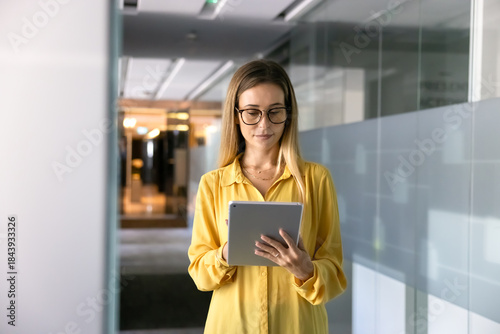 Serious young businesswoman in glasses typing on tablet pc in office corridor, working on Internet project, using digital technology for online business process management, browsing social media