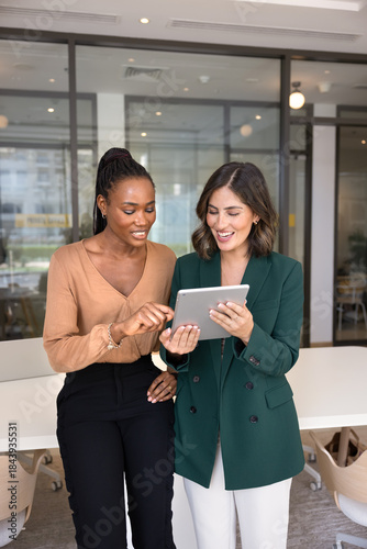 Positive multiethnic female coworkers discussing task on tablet, standing together in office space. Latin professional woman asking African colleague for help, advice, feedback, showing screen