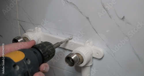 Professional plumbers use a power drill to drill a hole in a ceramic wall tile to install a new faucet during a bathroom renovation. A man installs a new faucet in the bathroom.