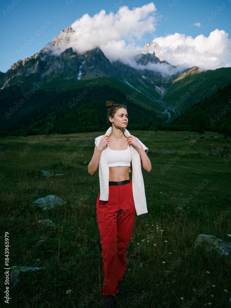 Fototapeta premium Woman in red pants and white top stands in alpine meadow with towering mountains, clouds and blue sky, enjoying outdoor scenery and nature tranquility.