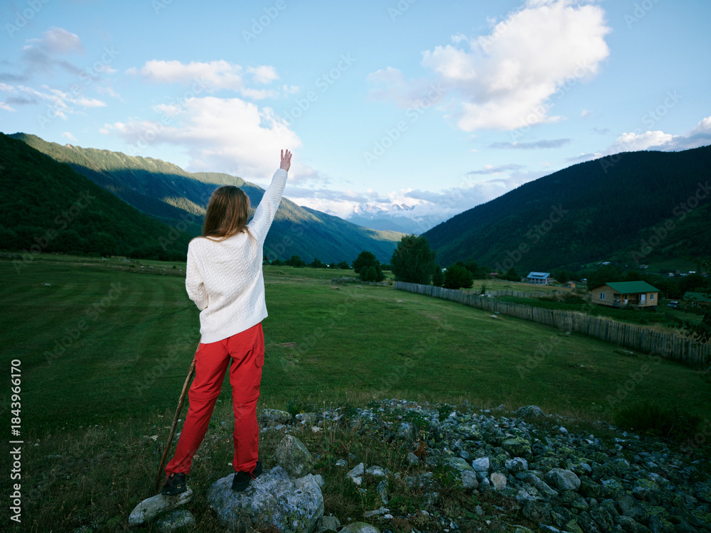 Fototapeta premium Woman standing on a rocky foreground, raising her hand toward a bright blue sky, with distant mountains and a green valley, conveying exploration, freedom, and outdoor adventure