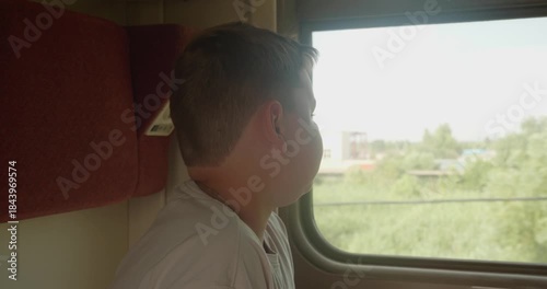 A pensive boy traveling on a train, admiring the green landscape passing by. The child enjoys the train ride, sitting in a comfortable passenger seat and looking out the window of the moving train.