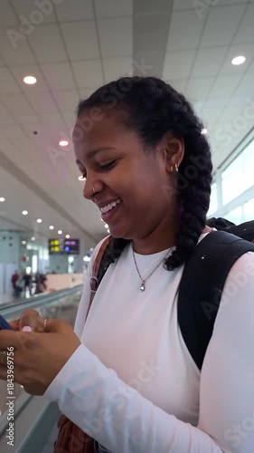 Vertical. African millennial woman checking her smartphone while pushing a luggage trolley in a bright airport terminal. Glass walkway and signage suggest boarding time, travel mood and anticipation.