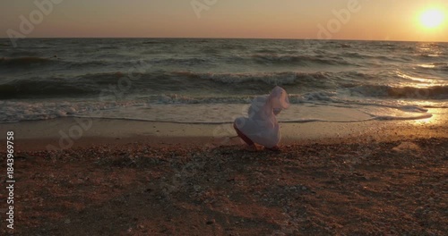 The child sits on the seashore and draws a stick on the sand, while the waves spray during excellent sunset, embodying peaceful children's vacations. The child plays with sea waves during sunset.