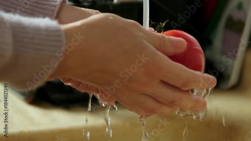 Slow motion video of a woman washing one fresh tomato under running water at home kitchen sink
