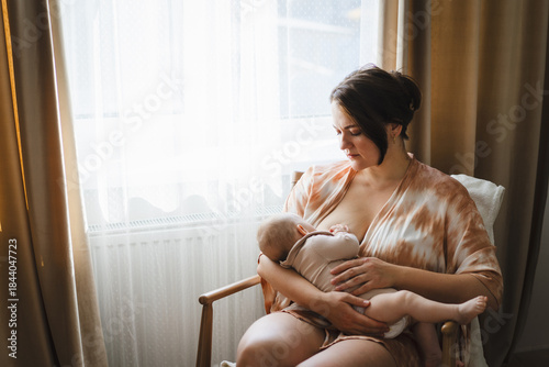 A mother sits in a wooden chair, gently breastfeeding her baby in a warm and softly lit room. Sunlight filters through the curtains, creating a peaceful atmosphere.
