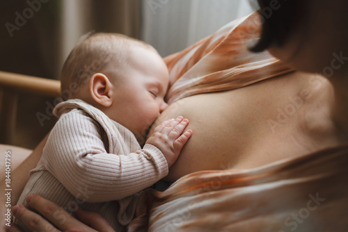A mother sits in a wooden chair, gently breastfeeding her baby in a warm and softly lit room. Sunlight filters through the curtains, creating a peaceful atmosphere.