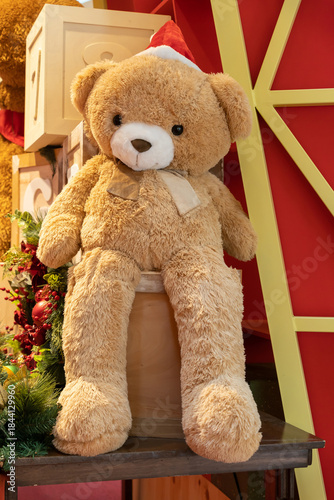 A soft tan teddy bear wearing a red Santa hat sits among festive Christmas decor, gift boxes, and holiday