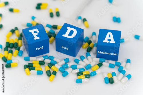 Three blue blocks labeled ‘FDA’ (Food and Drug Administration) are positioned amid a dispersed collection of pharmaceutical capsules, symbolizing regulatory oversight