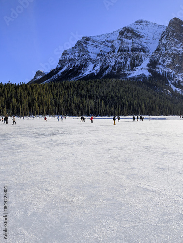 Visitors ice skate on a natural ice rink - frozen Lake Louise in winter. Winter landscape in snow-capped mountains passes by a frozen lake leading to the town - Lake Louise, Alberta, Canada.